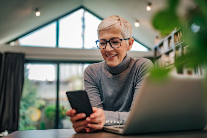 Close-up portrait of a happy senior woman looking at smart phone while sitting at desk with laptop.
