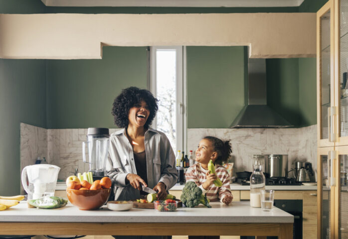 An African-American Single Mother Preparing Vegan Lunch In The Kitchen And Smiling With Her Little Daughter